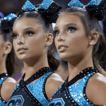 Three cheerleaders in matching blue and black sequined uniforms with large blue bow headbands, standing in an arena.